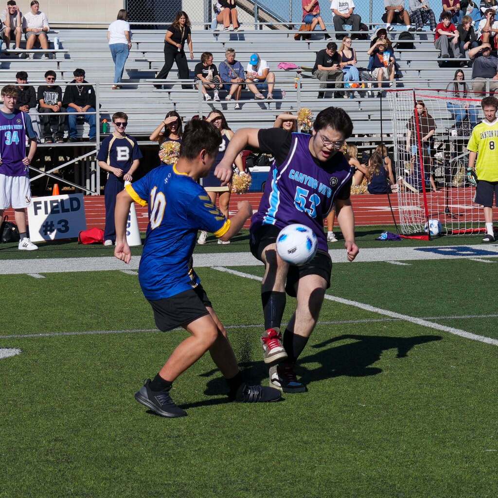 Image 8: Two Unified soccer athletes in blue and purple jerseys battling for possession of a soccer ball on the field in front of filled stadium bleachers.