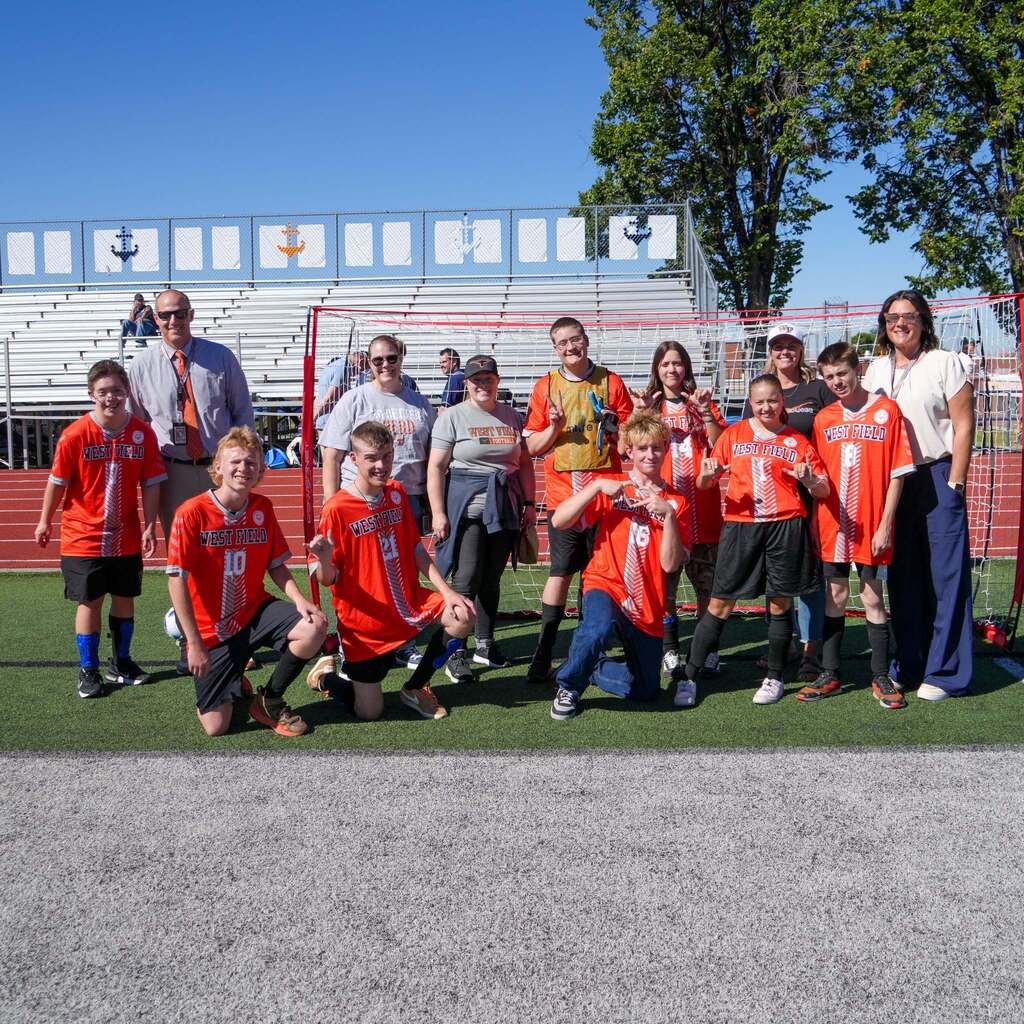 Image 2: Westfield High Unified Sports players and coaches standing together in front of a soccer goal frame on the turf field.