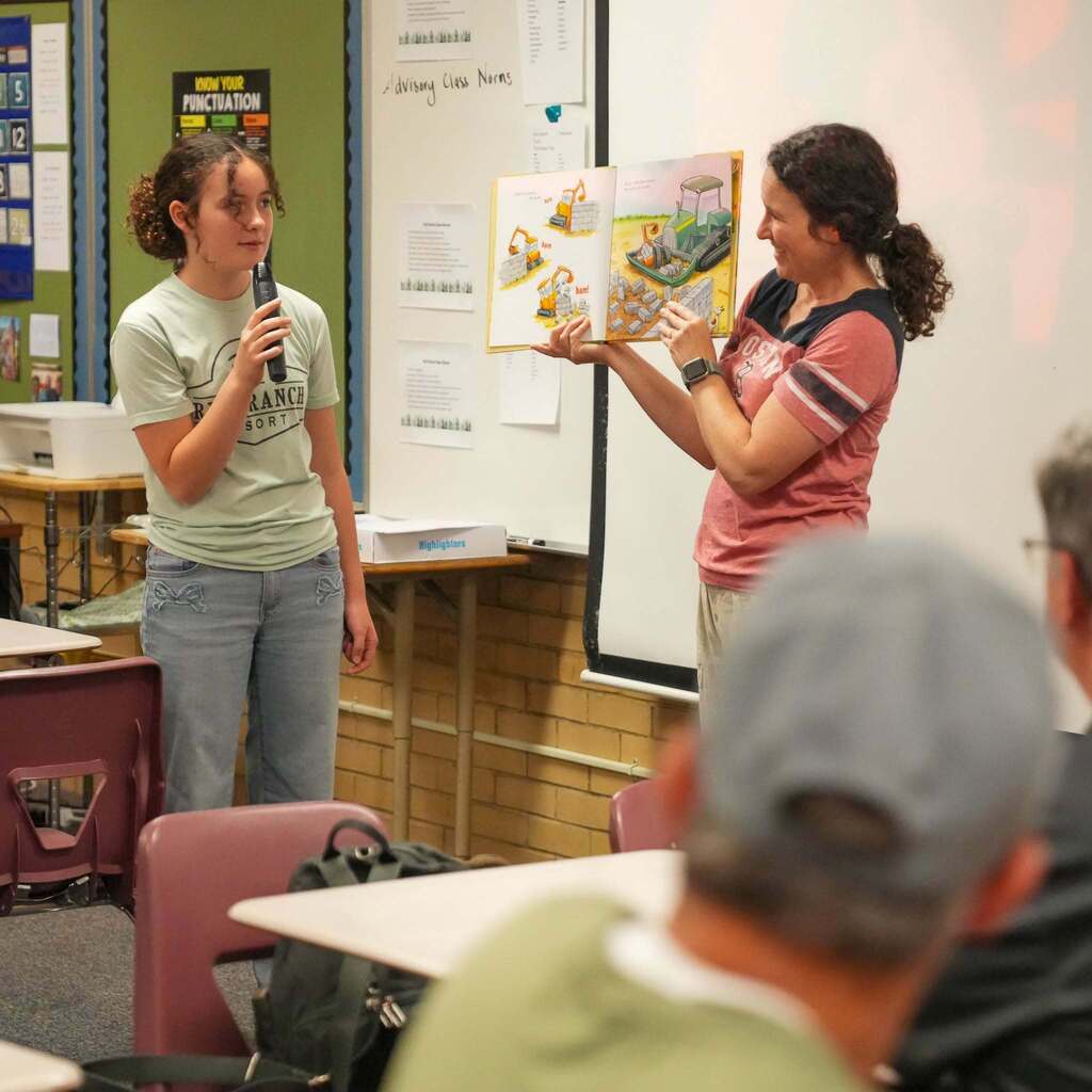 Image 2: A student holding a microphone while an educator displays open pages of a colorful children's book to a classroom audience.
