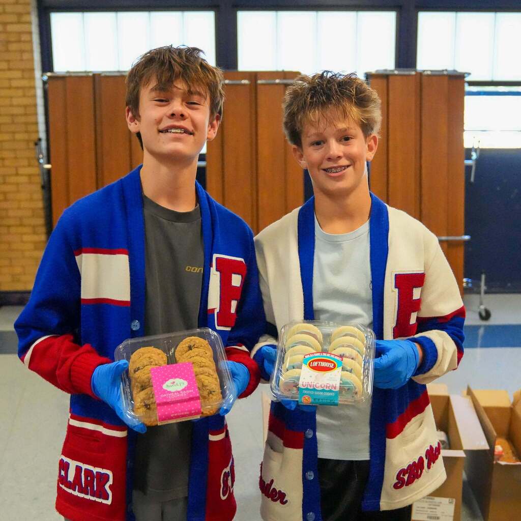 Image 6: Two smiling male students wearing school letterman cardigans and blue gloves holding containers of cookies to serve at the event.