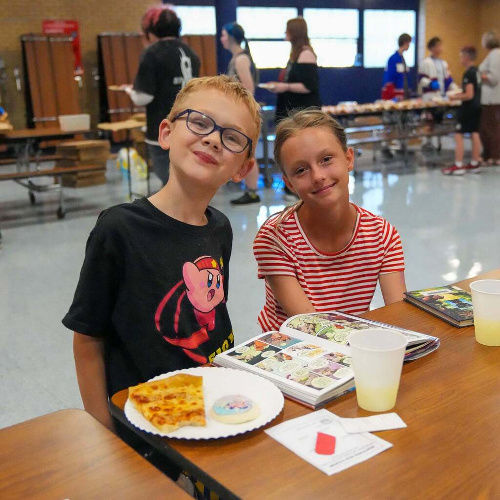 Image 7: Two young students seated at a cafeteria table enjoying pizza and reading a graphic novel together.