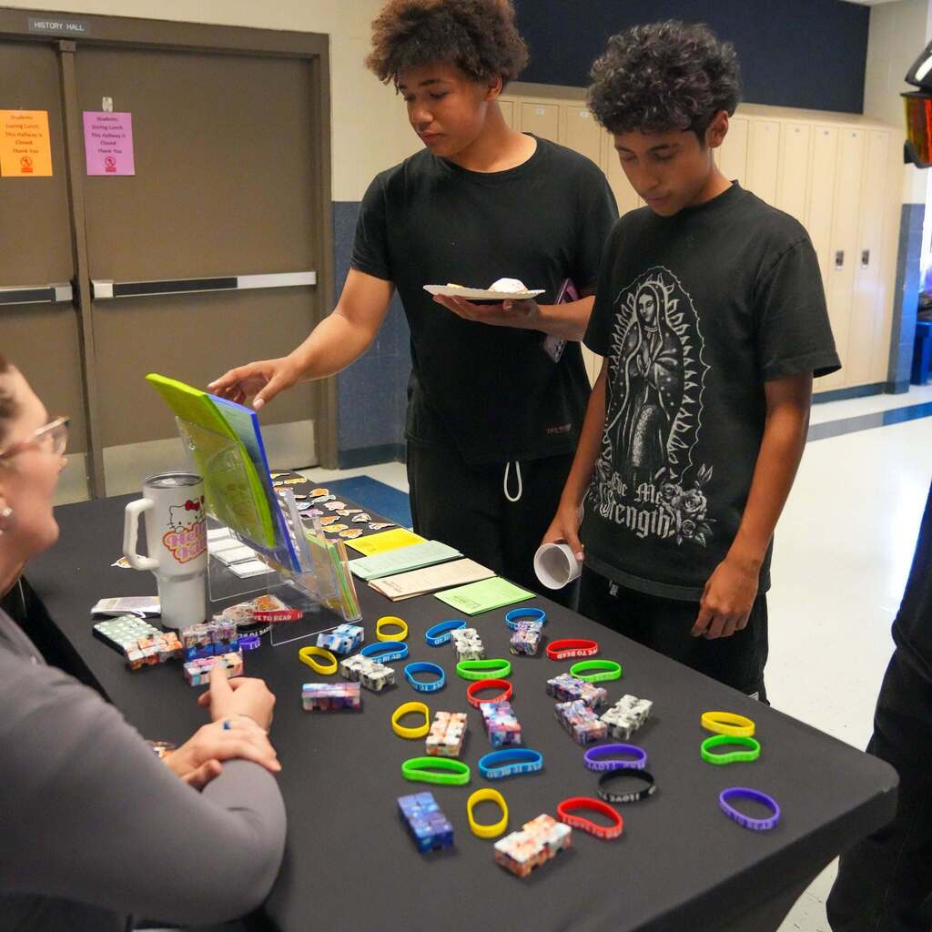 Image 8: Two male students browsing colorful wristbands and fidget toys displayed on an information table hosted by a school staff member.