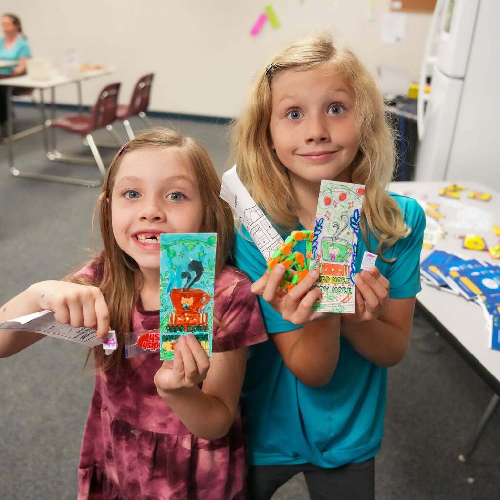 Image 4: Two young students proudly showing off their hand-colored bookmarks and small prize items during Reading Night activities.