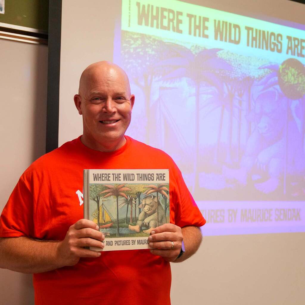 Image 10: A smiling educator in a bright red shirt holding up a copy of Where the Wild Things Are in front of a screen projecting the book's cover artwork.
