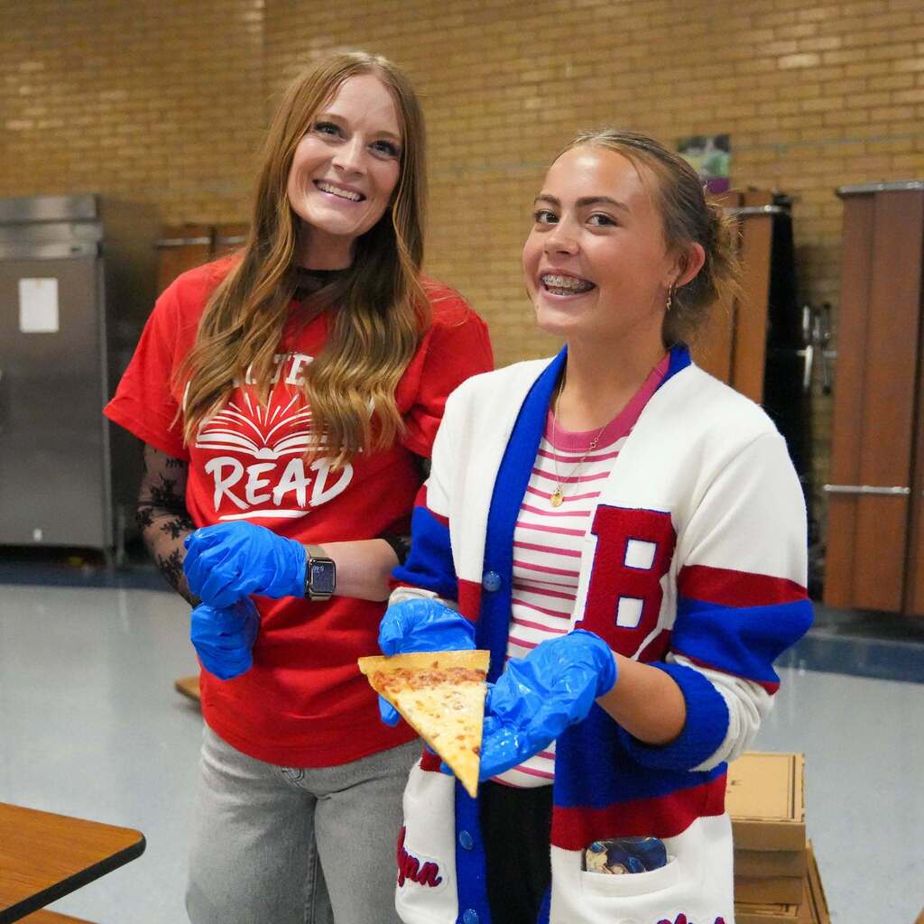 Image 5: A staff member and a student in a school cardigan wearing blue protective gloves while holding up a slice of pizza to serve attendees.