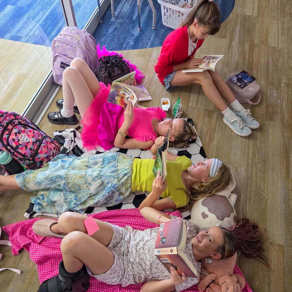 Image 7: Four female students lying and sitting together on blankets on the floor near a bright window, deeply engrossed in reading their books.
