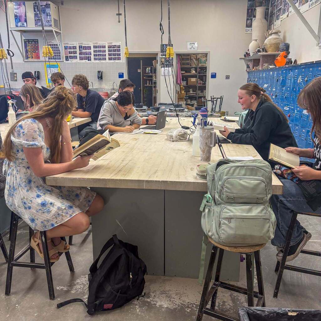 Image 5: Students sitting on high stools at a large table inside an art studio room while reading books during designated reading time.