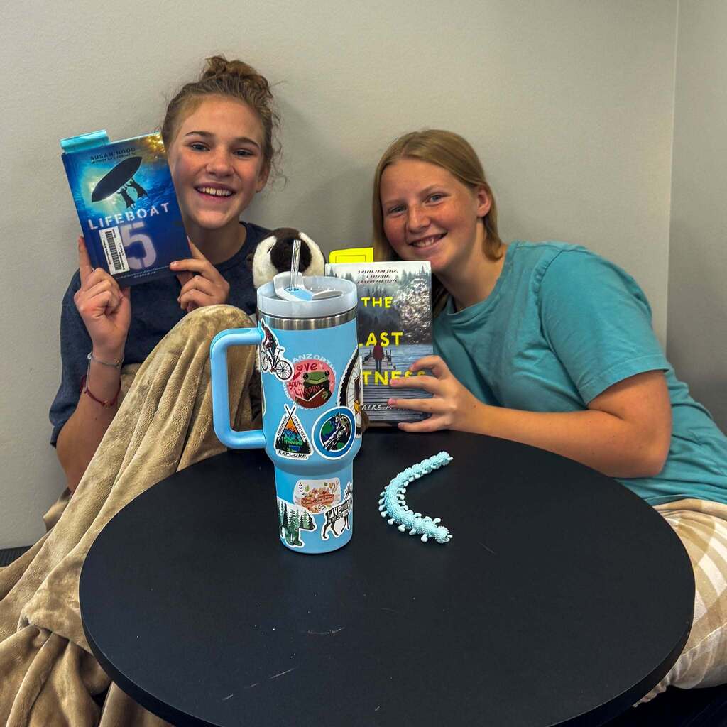 Image 4: Two students smiling side-by-side at a small table, proudly holding up book covers alongside a large decorated water bottle and a fidget toy.