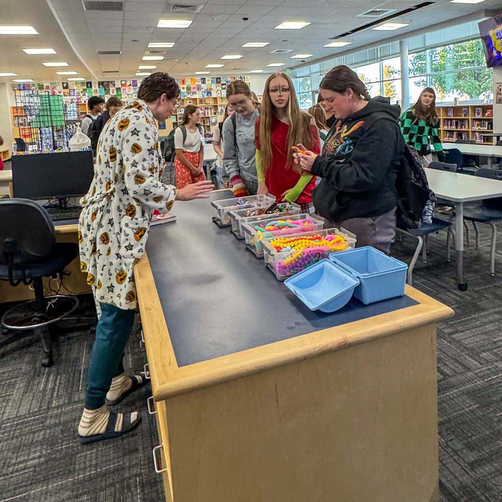 Image 3: Students gathered around a counter in the school library choosing colorful craft supplies and fidget items with a staff member dressed in a Halloween robe.