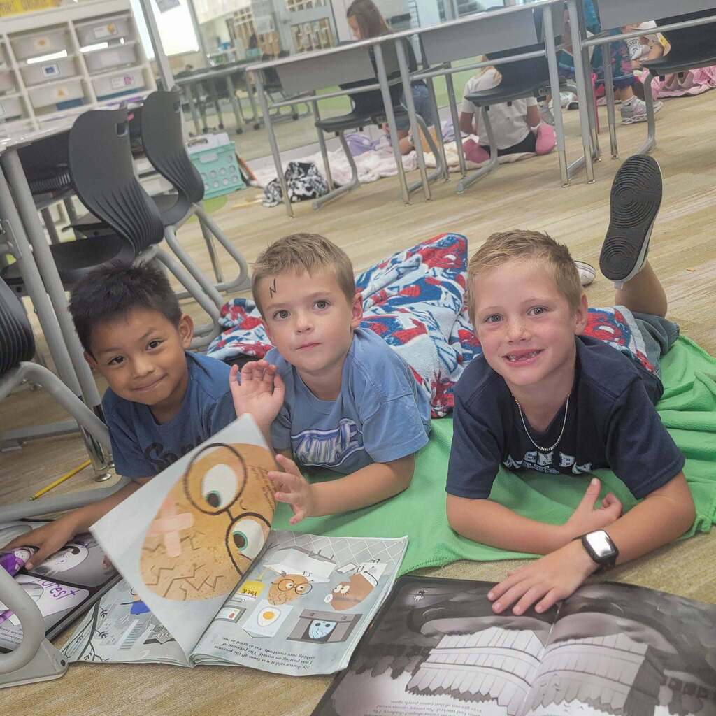Image 10: Three young boys lying on their stomachs on a soft green blanket on the classroom floor, smiling as they look at picture books open in front of them.
