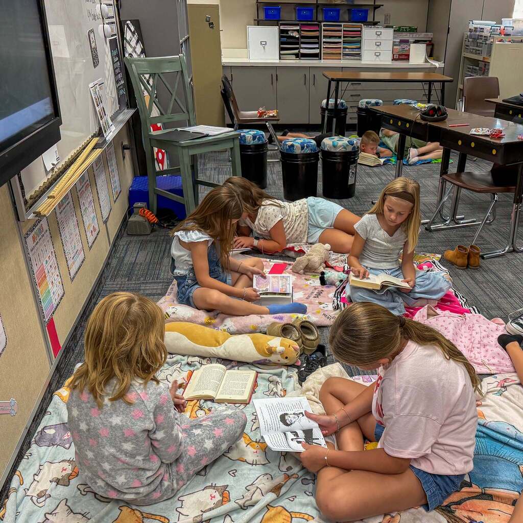 Image 1: Five elementary school students wearing comfortable clothes and pajamas lounging on blankets across a classroom floor while reading books together.
