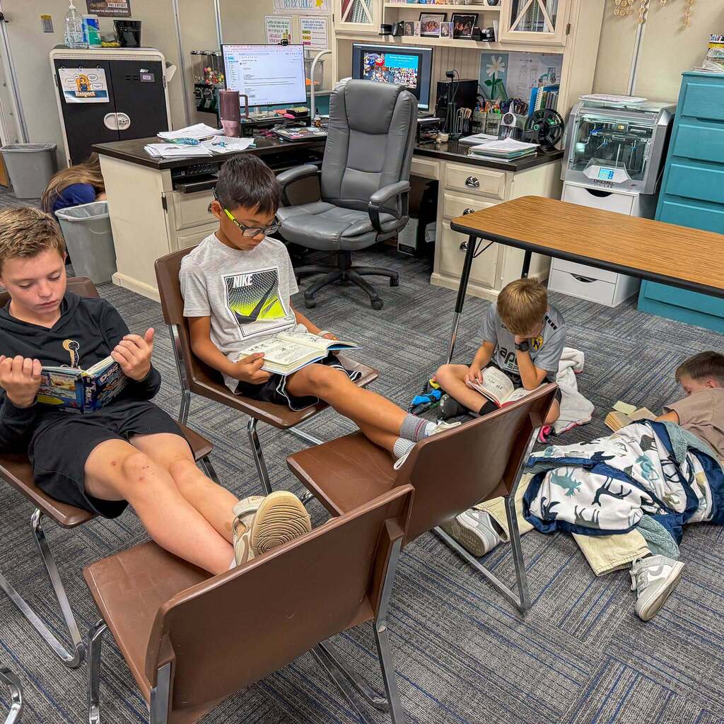 Image 6: Four young students finding cozy spots—on chairs with their feet resting on other chairs, on the floor with blankets, or under desk tables—to read books in a classroom.