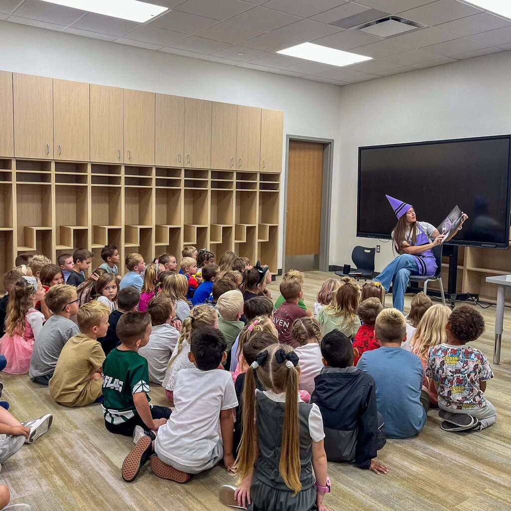 Image 8: A guest reader wearing a purple crayon hat reading a storybook aloud to a large class of elementary students sitting attentively on the floor.