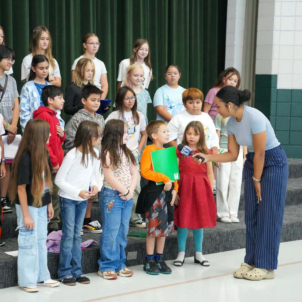 Image 9: A FOX 13 reporter leaning down with a microphone to interview a young student standing on stage with a group of classmates.