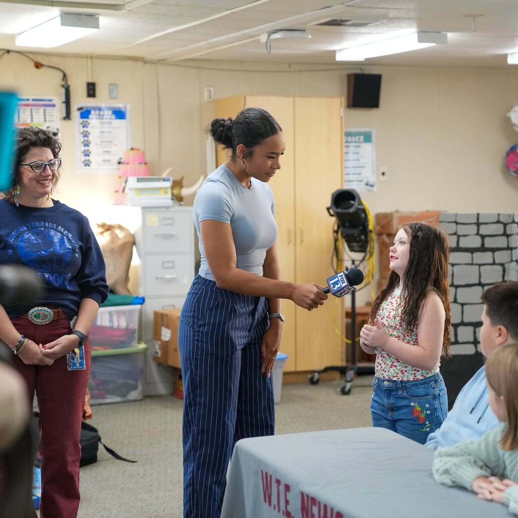 Image 5: A news reporter holding a microphone up to a young student speaking in a classroom while a teacher looks on and smiles.