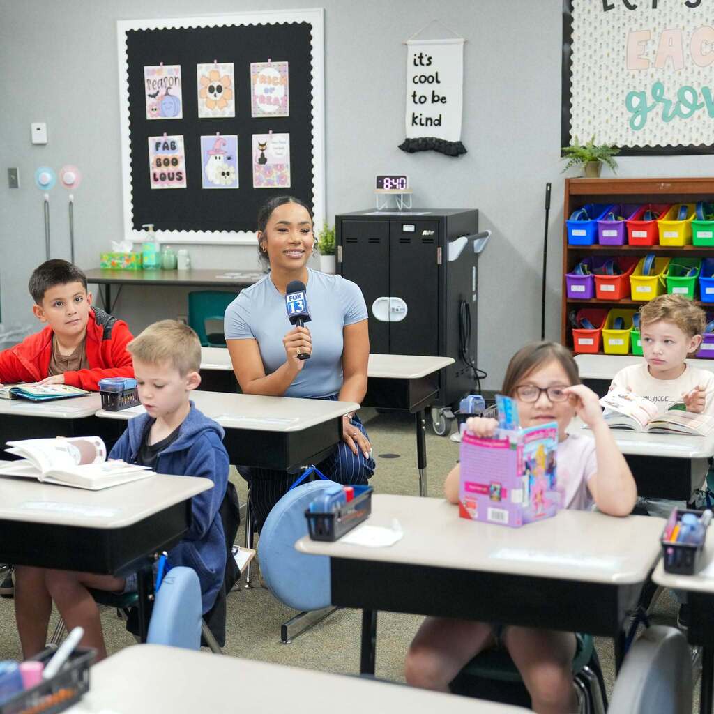 Image 6: A FOX 13 reporter holding a microphone while sitting amongst elementary students at their classroom desks during reading time.