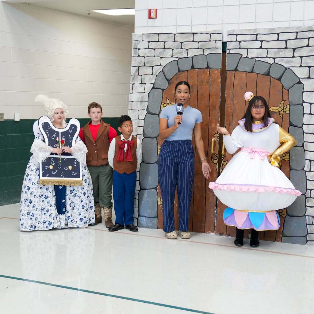 Image 8: A news reporter standing in front of a painted castle doorway backdrop with elementary students wearing theater costumes for a school play.