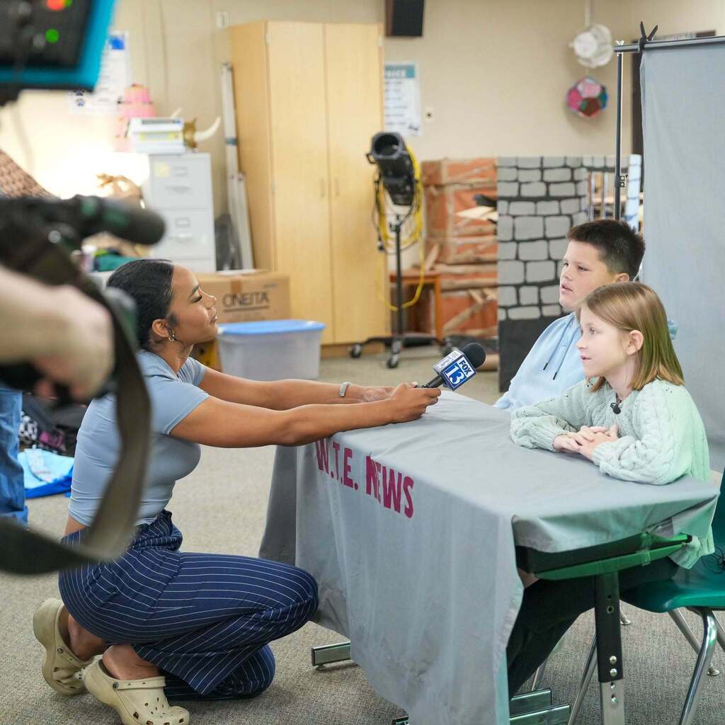 Image 4: A FOX 13 news reporter kneeling down with a microphone at a news desk set up in a classroom to interview two elementary student news anchors.