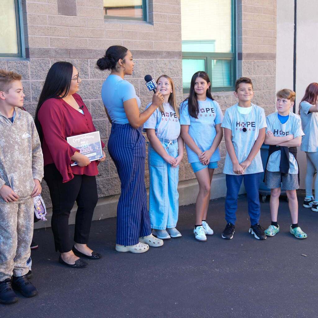 Image 10: A news reporter interviewing members of the school's Hope Squad and staff members standing outside against the brick building wall.