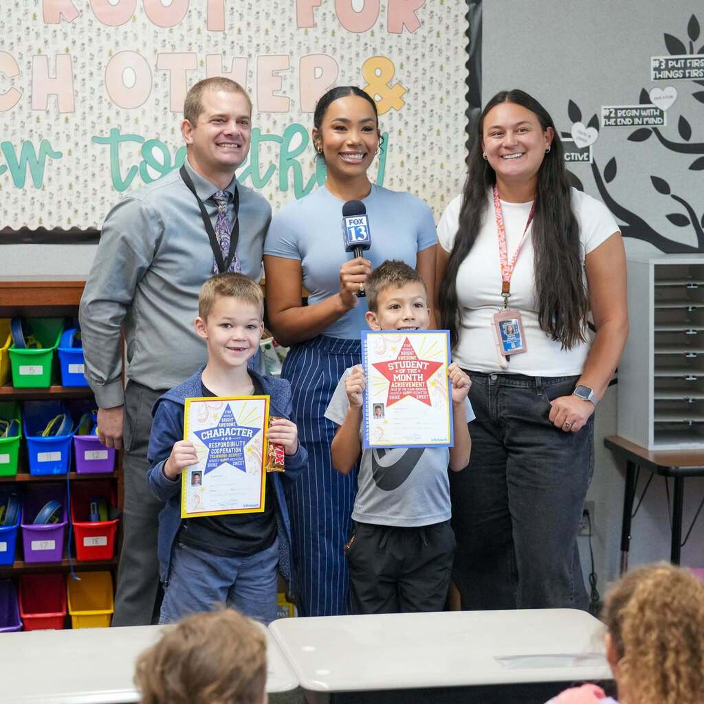 Image 2: A FOX 13 news reporter holding a microphone and standing with school administrators and a teacher as two young students proudly hold up their Student of the Month certificates.