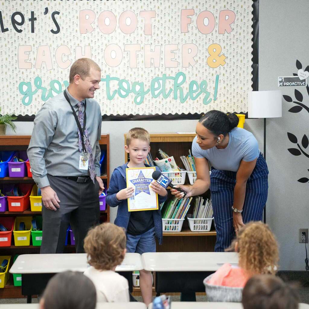 Image 7: A news reporter extending a microphone to a young student holding an award certificate while a smiling school administrator stands beside them.