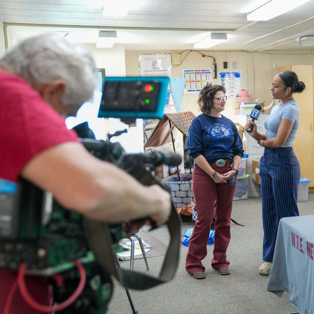 Image 1: A news videographer capturing a broadcast camera shot of a FOX 13 reporter interviewing a school staff member wearing a Washington Terrace Elementary Bulldogs sweatshirt.