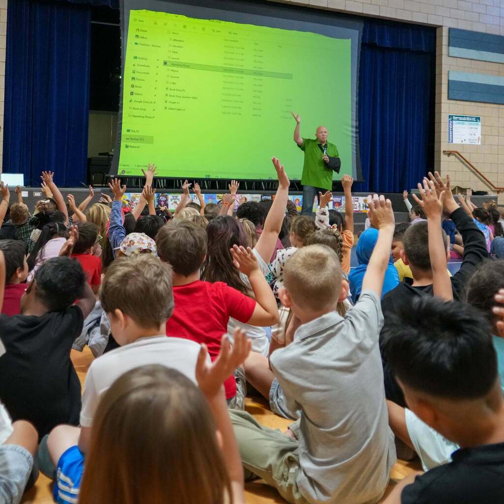 Image 7: Elementary students sitting on the gymnasium floor raising their hands eagerly to ask questions during an author assembly.