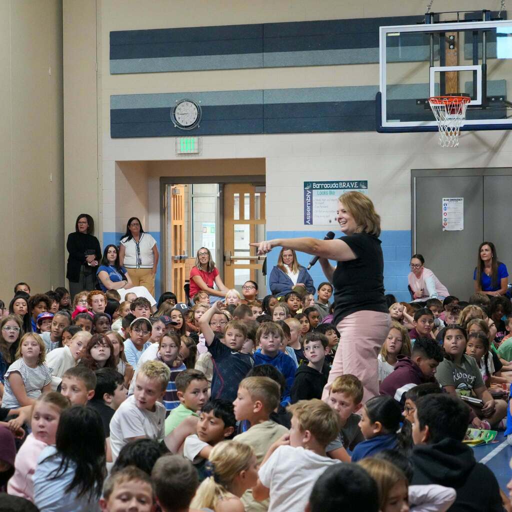 Image 2: Visiting author Jennifer A. Nielsen speaking into a microphone and gesturing toward the audience during a lively school assembly in the gymnasium.