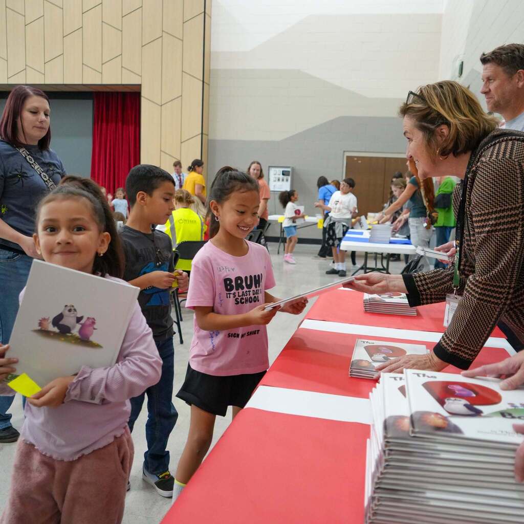 Image 4: District Superintendent Dr. Gina Butters and staff handing out new books to smiling elementary students standing in line.