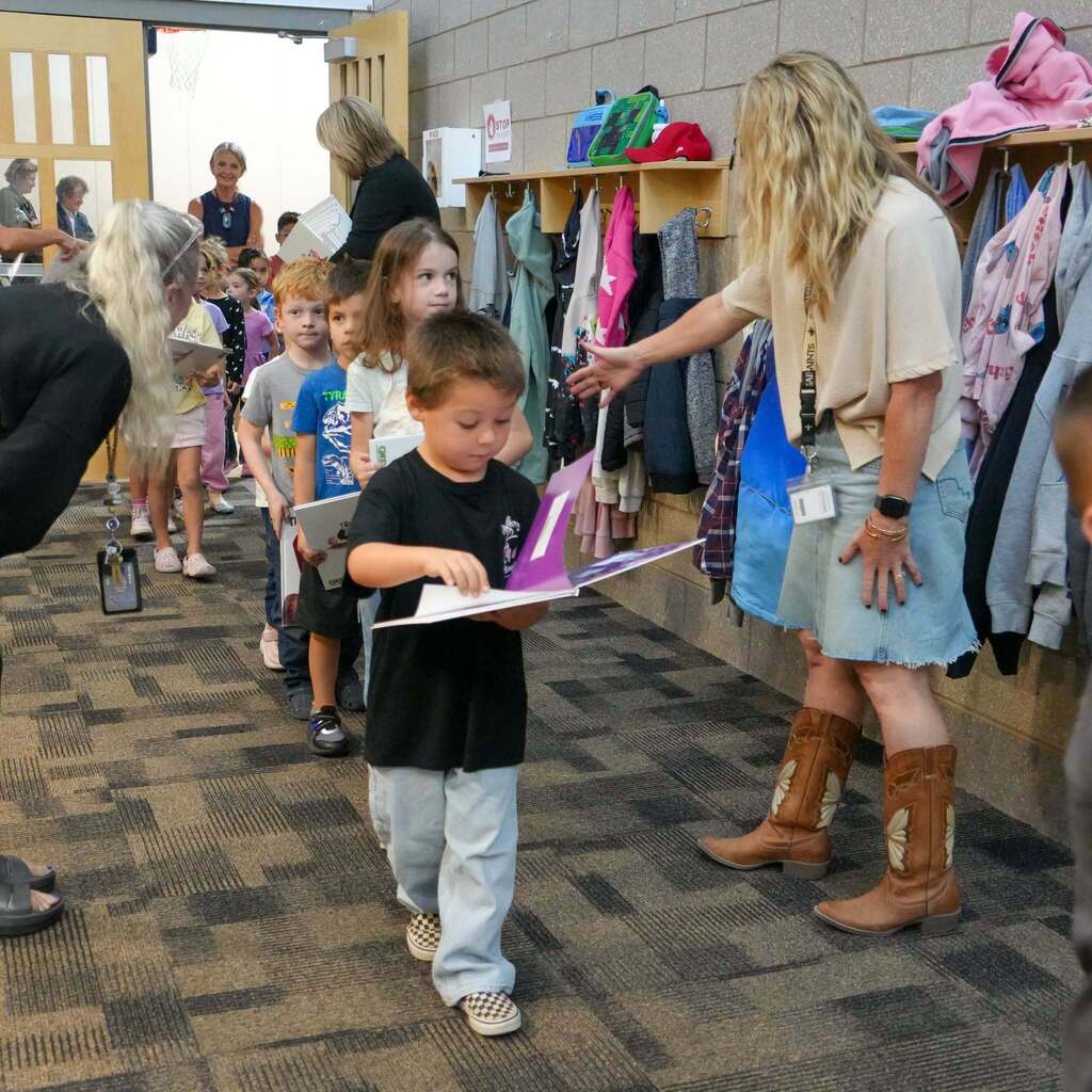 Image 10: A teacher wearing cowboy boots directing a line of young elementary students carrying their new books out of the classroom.
