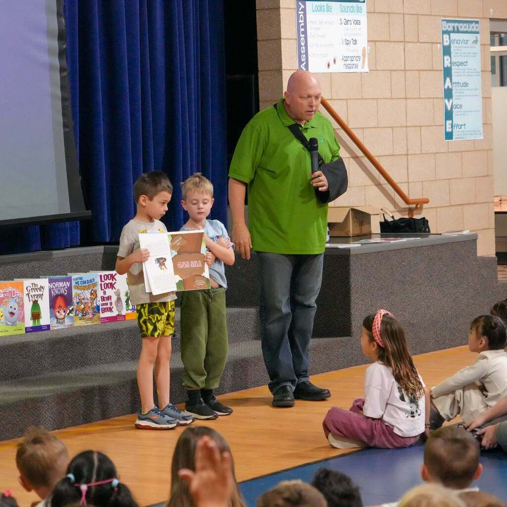 Image 9: An author with his arm in a sling guiding two young students on stage as they hold open a picture book for the audience.