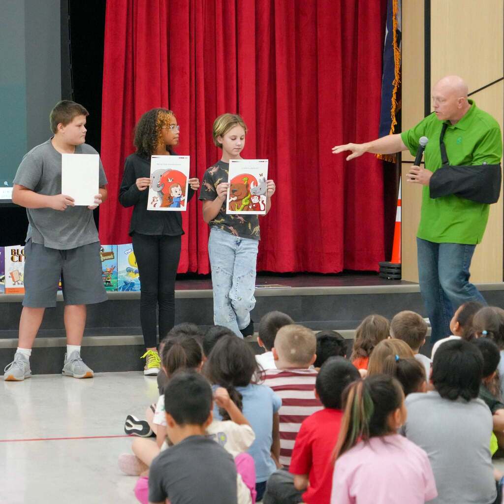 Image 3: A visiting author with a arm sling presenting on stage alongside three elementary students holding up illustrated story pages.