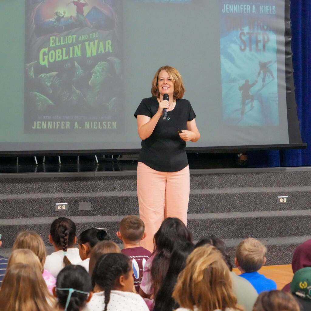 Image 5: Author Jennifer A. Nielsen presenting on stage in front of a screen displaying her book covers to a gymnasium full of seated students.
