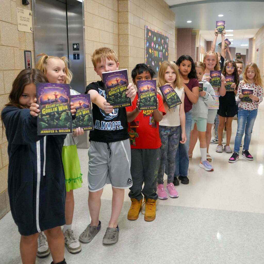 Image 1: A line of elementary students standing in a hallway, excitedly holding up copies of the book Elliot and the Goblin War by Jennifer A. Nielsen.