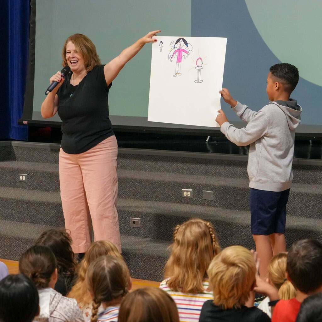 Image 6: Author Jennifer A. Nielsen laughing and holding up a student's artwork on poster paper on stage next to a student assistant.