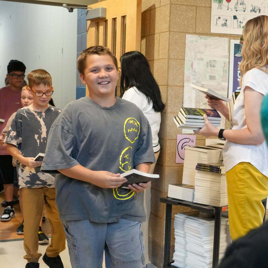 Image 8: A smiling elementary student walking down the hallway carrying a book after receiving it from stacks set up on a mobile cart.