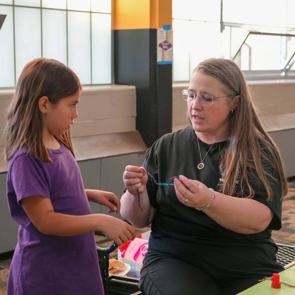 Image 4: A staff member sitting and demonstrating how to weave a colorful string bracelet to a young student inside the school gymnasium.