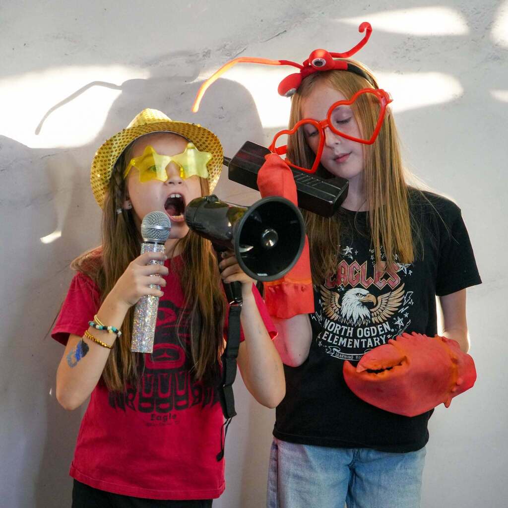 Image 3: Two young elementary students wearing fun dress-up accessories—including star-shaped sunglasses, a sequined hat, and lobster claws—speaking and chanting into a microphone and megaphone.