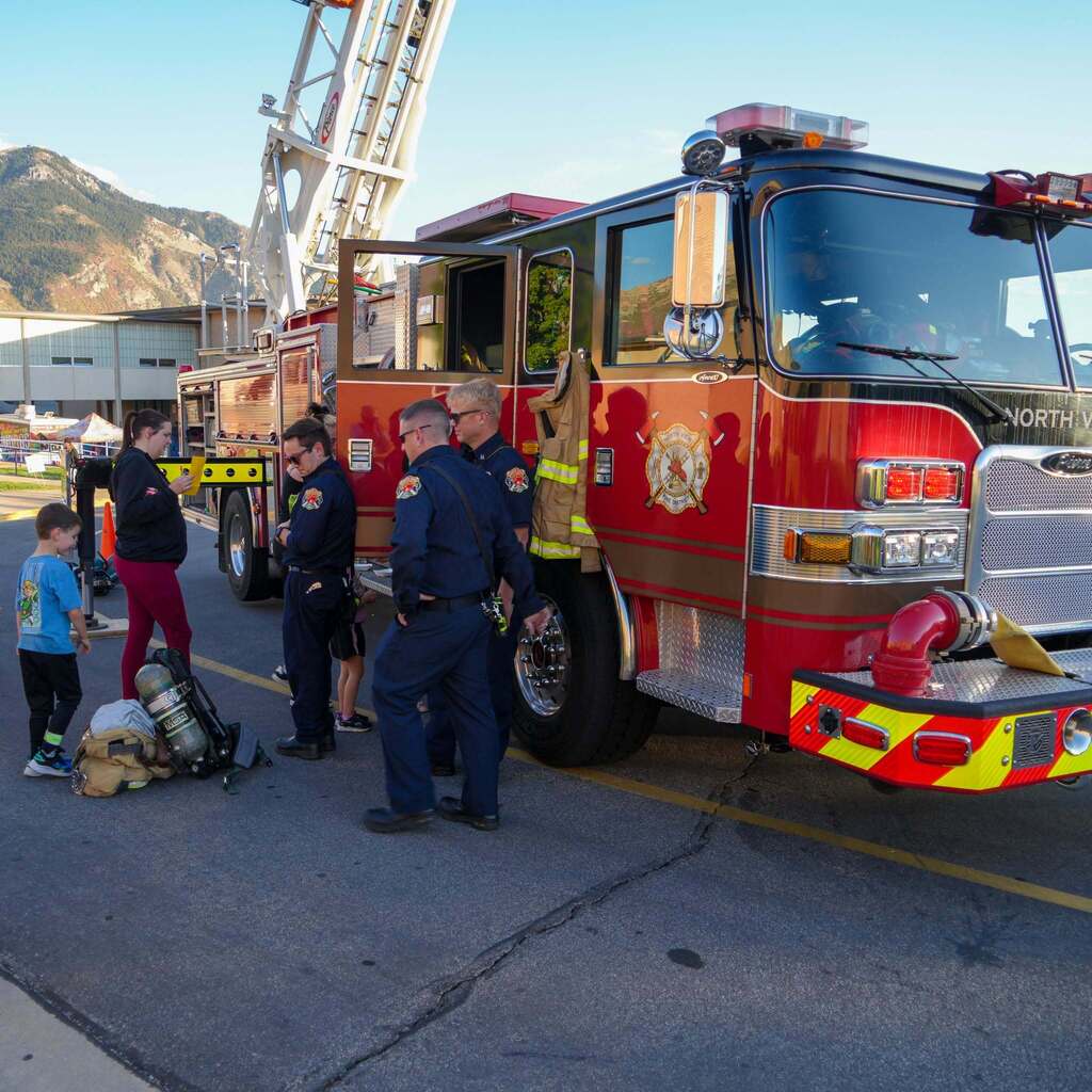 Image 1: A group of firefighters standing outside a large North View Fire District ladder truck parked at a school community event while a young student and parent look on.