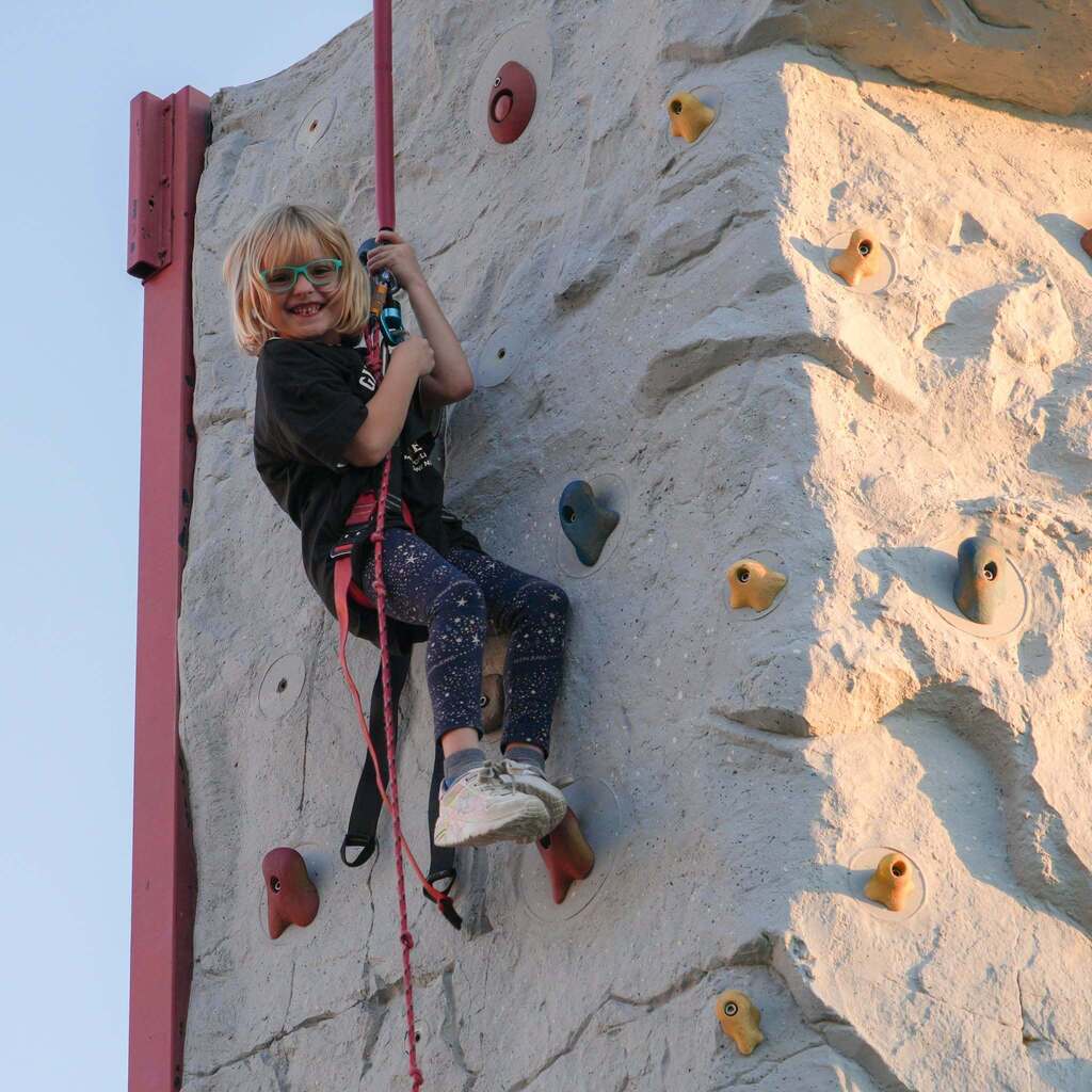 Image 9: A smiling young girl with glasses climbing up an outdoor artificial rock wall while wearing a safety harness and helmet strap.