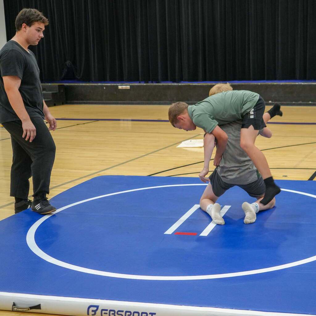 Image 7: Two young students practicing wrestling moves on a blue padded mat inside the gymnasium under the supervision of an instructor.