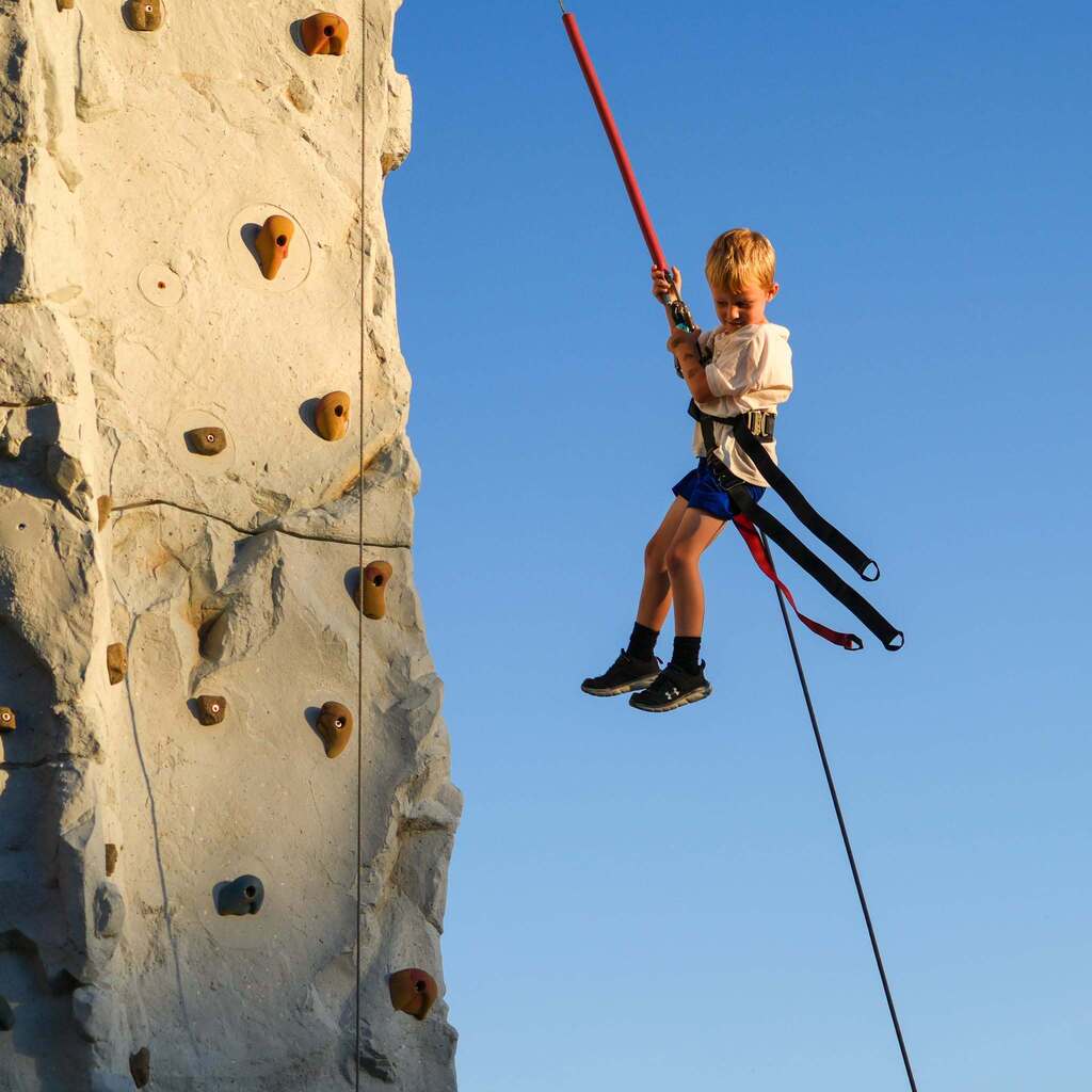 Image 8: A young student secured in a safety harness lowering down from an outdoor mobile climbing wall against a clear blue sky.