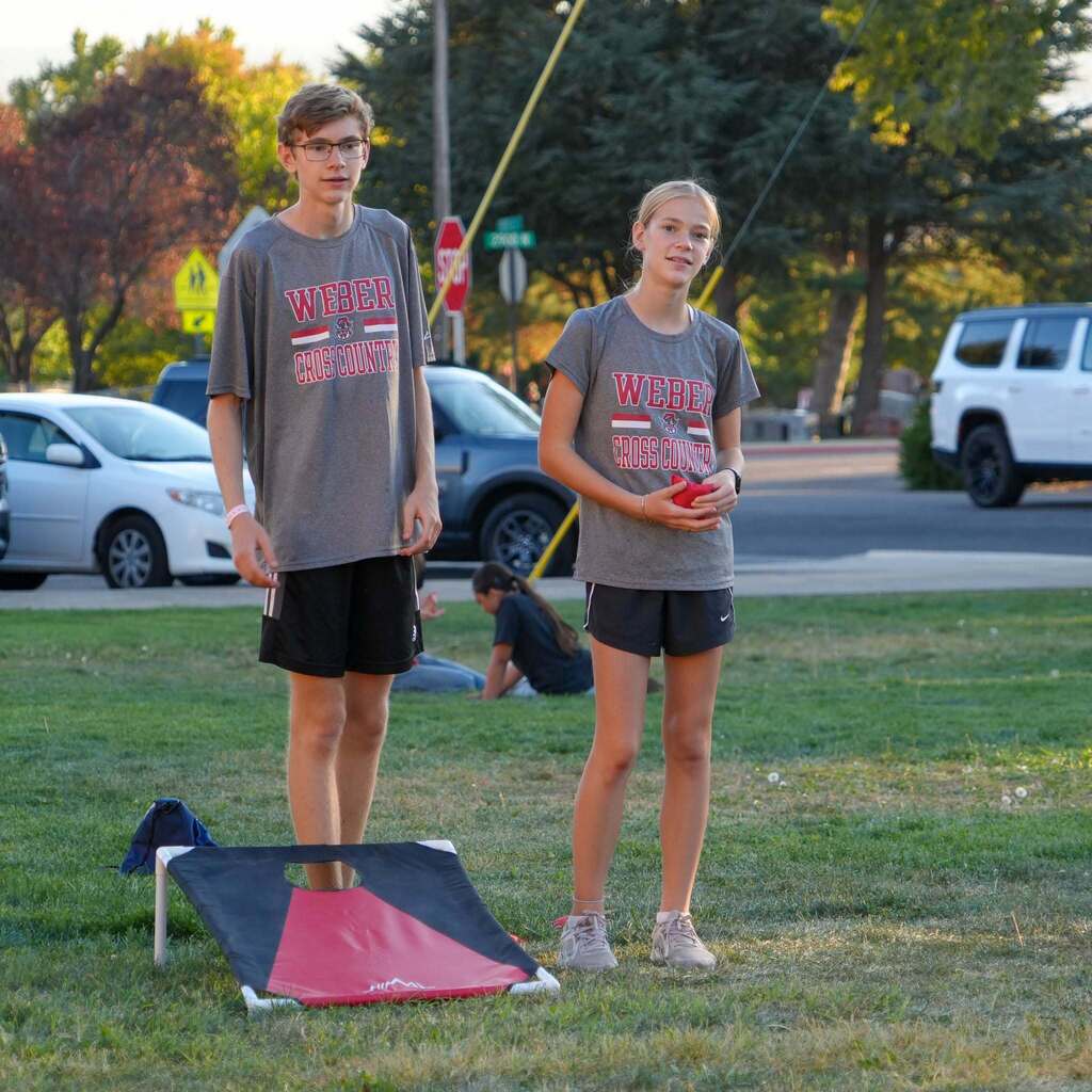 Image 2: Two student-athletes wearing grey Weber Cross Country t-shirts standing on a grassy field playing a game of cornhole.