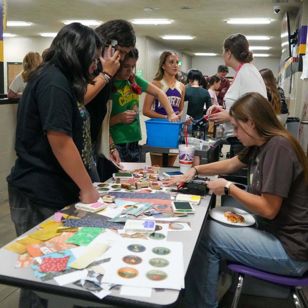 Image 5: Students gathered around a craft table in a school hallway selecting paper shapes and stamps to make custom buttons and bookmarks.