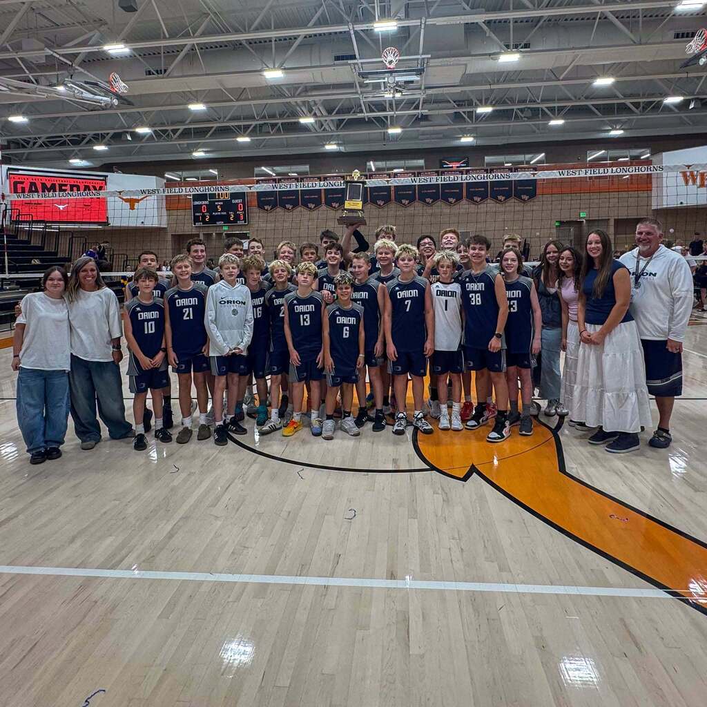 Image 1: The Orion Junior High boys volleyball team and coaching staff posing together on the court holding their championship trophy after winning the tournament.