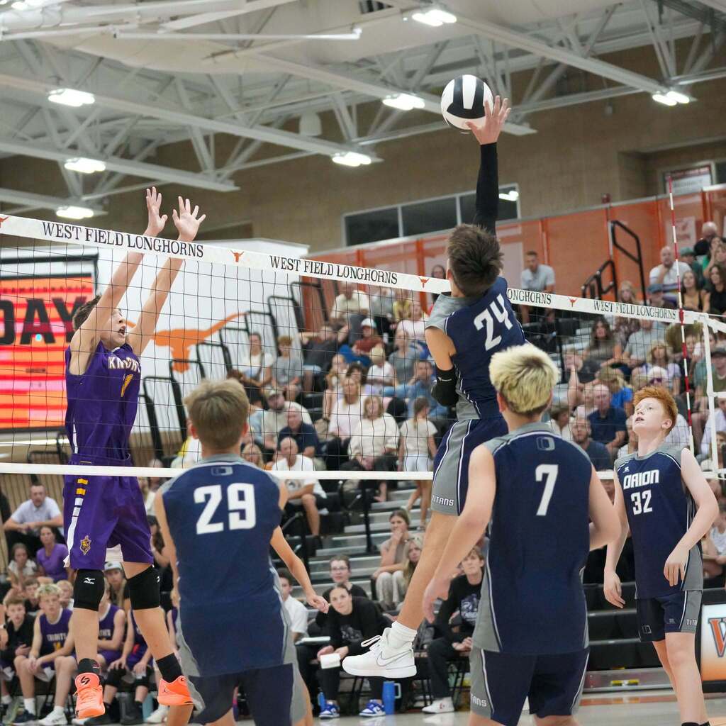Image 8: An Orion Junior High player soaring above the net to hit a spike while opposing defenders leap to block.