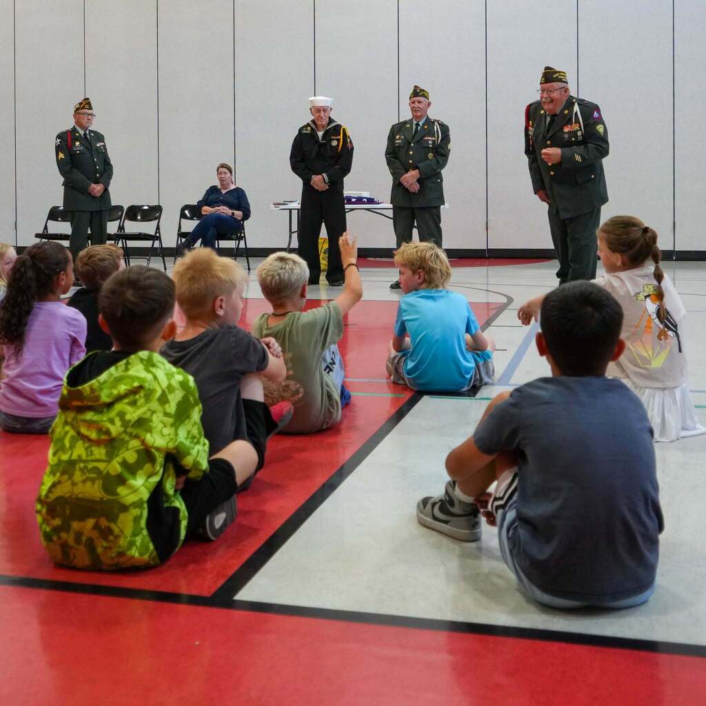 Image 2: Students sitting attentively on the gymnasium floor raising their hands as a panel of military veterans speaks to them.