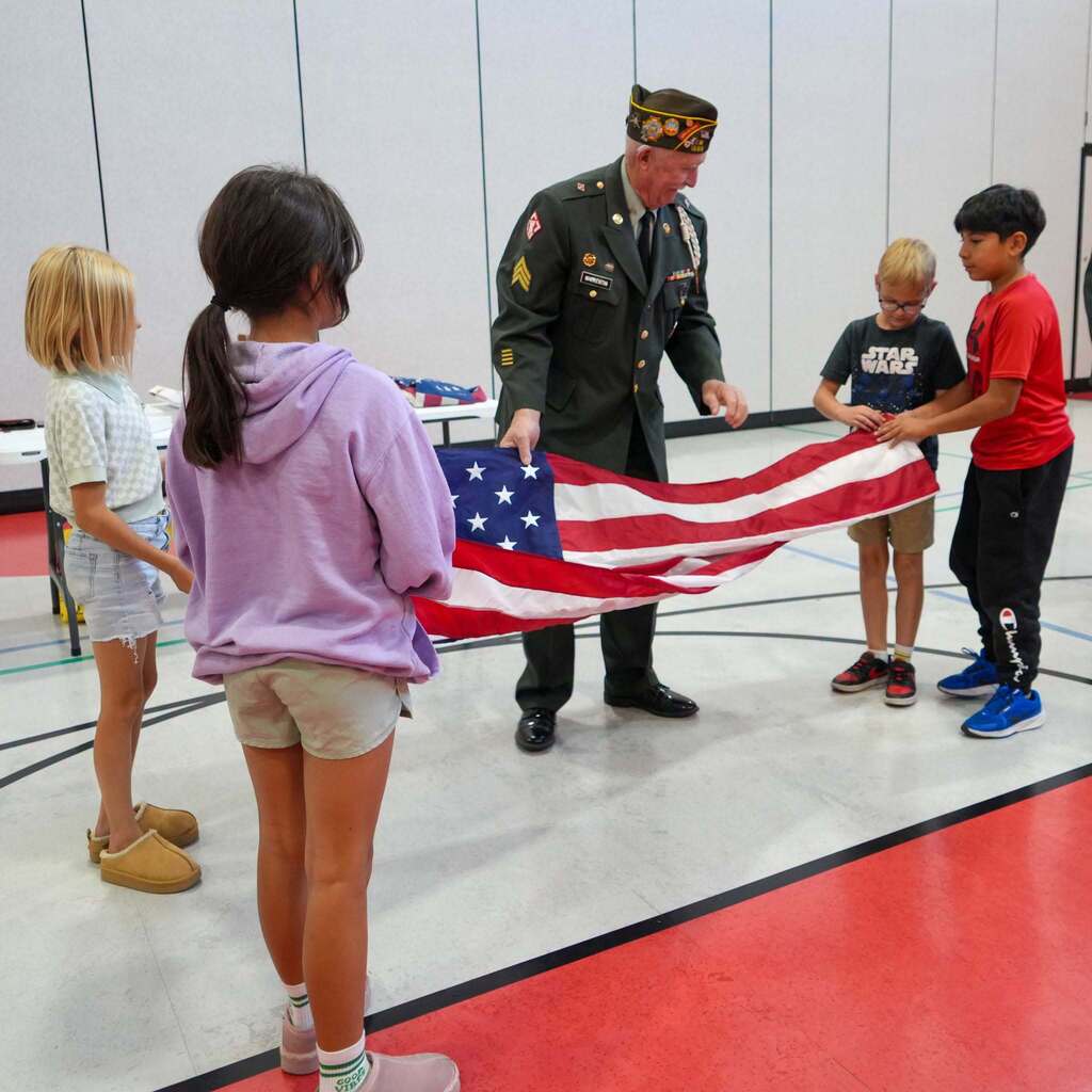 Image 5: A veteran assisting four students standing in a circle as they work together to pull the American flag taut for folding.