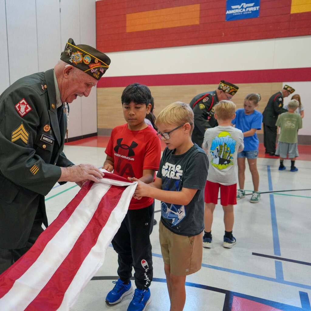 Image 1: A military veteran in uniform guiding two young students step-by-step as they learn how to fold an American flag inside the school gymnasium.