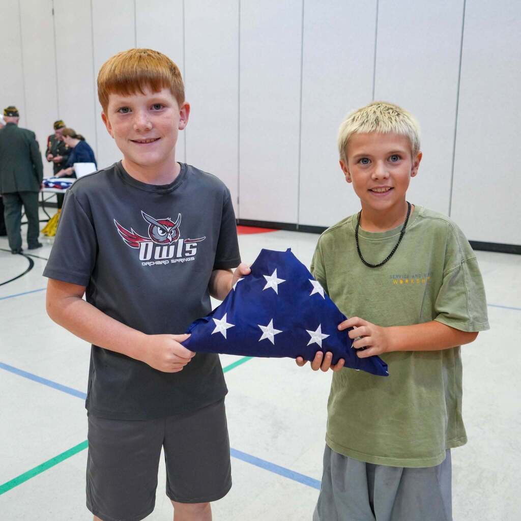 Image 10: Two elementary students smiling proudly as they hold up a perfectly folded triangular American flag together.
